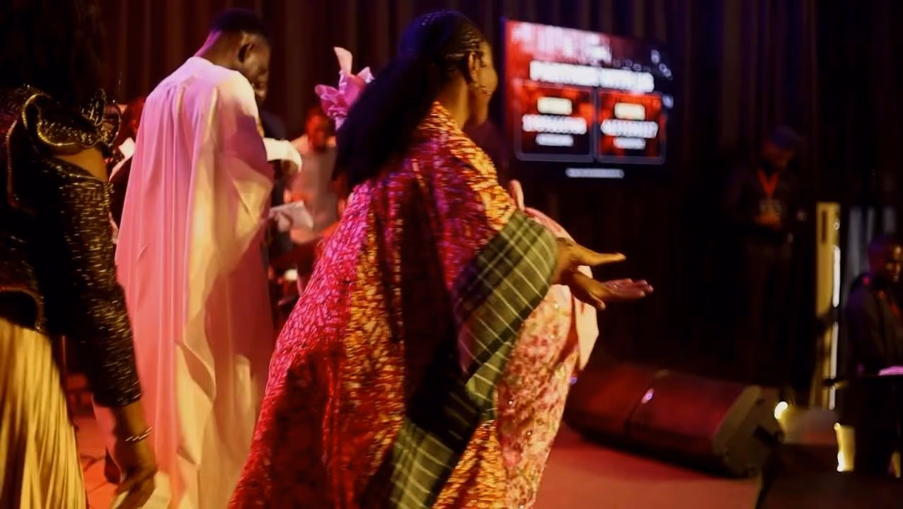 A woman in a patterned wrapper dances towards the stage beside a man in a white agbada