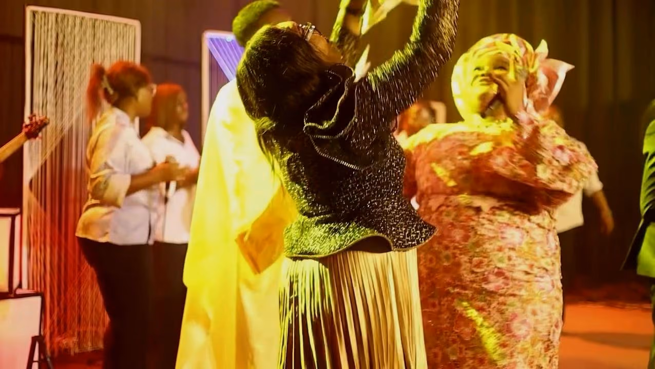 Two women in gele raise their hands in celebration on stage under warm yellow light