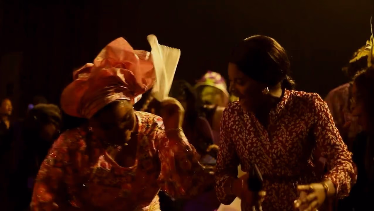 Two women dance together in the aisle, one in a pink gele waving a white handkerchief