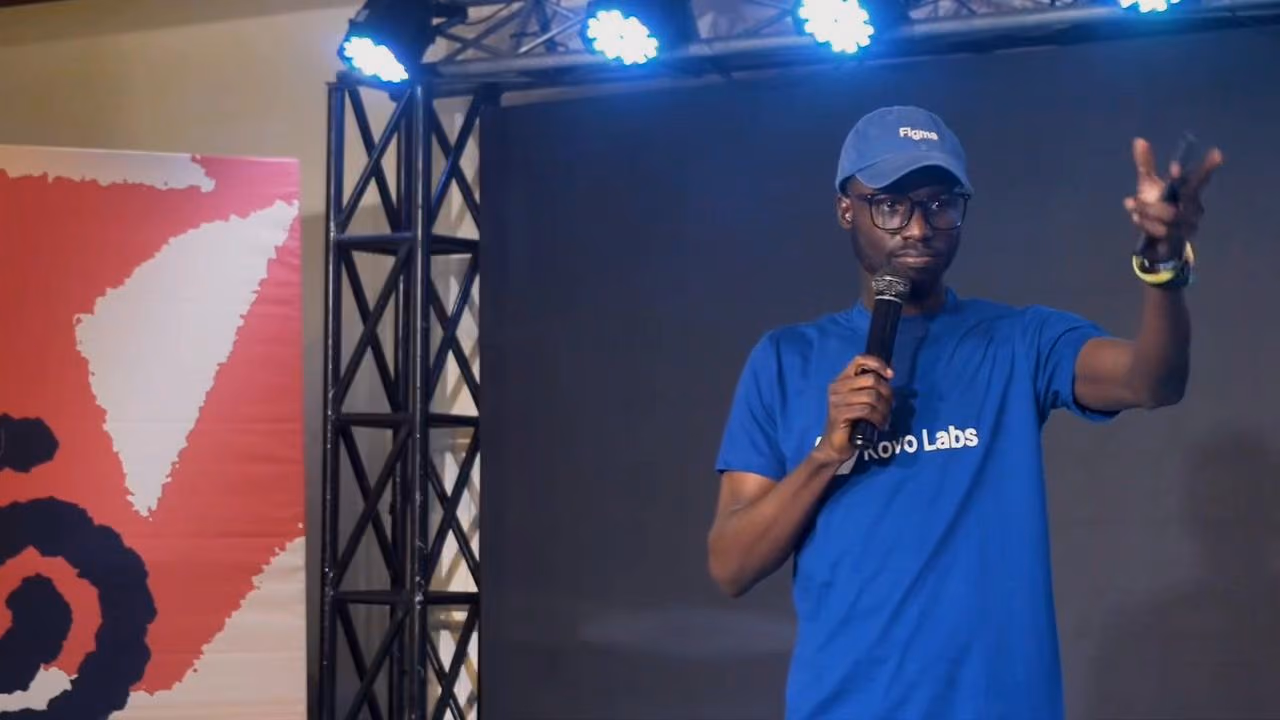 A speaker in a blue Figma cap and tee gestures to the audience under stage lights