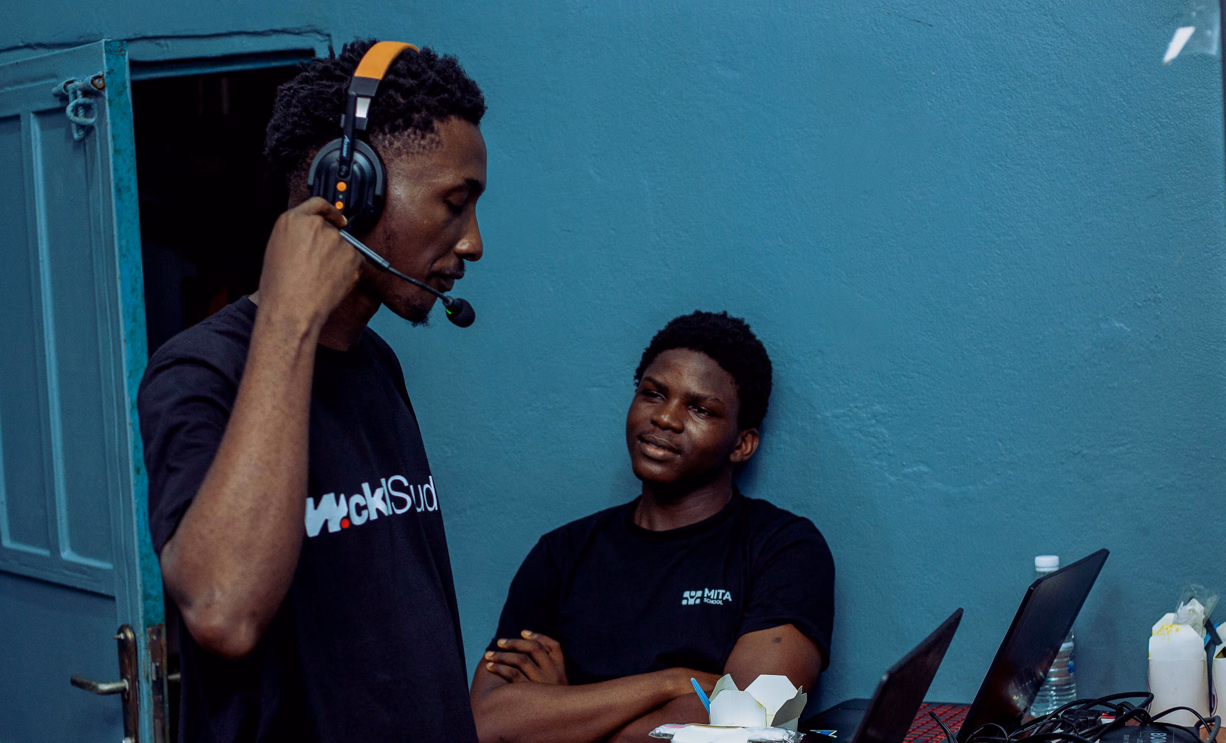 Operator on a headset standing beside a seated crew member in a small control room with laptops and cables