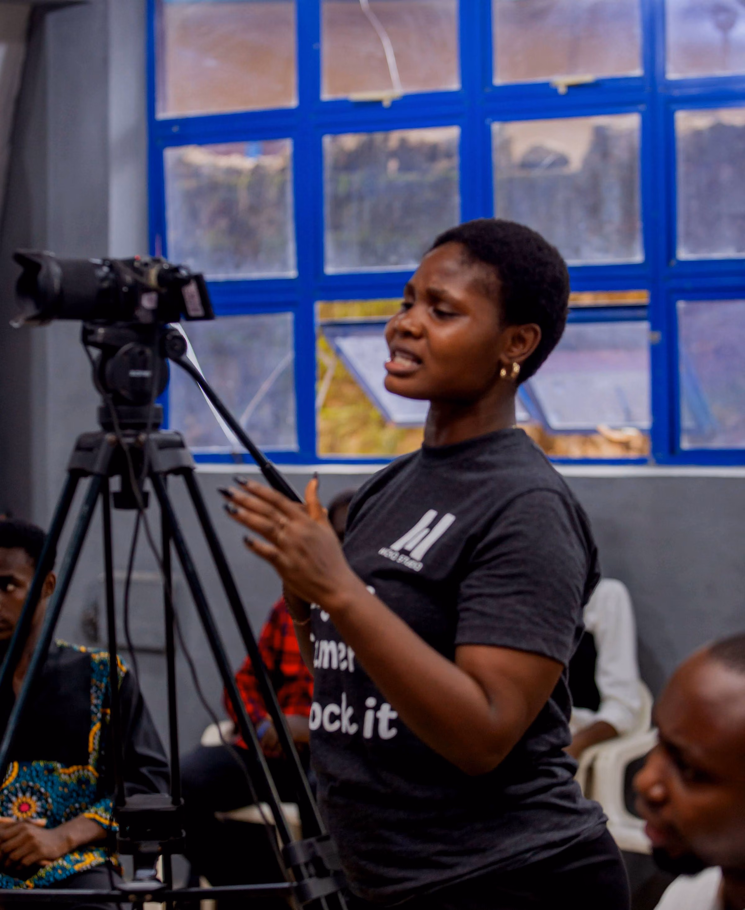 Camera operator in a grey tee on a tripod camera by a blue-framed window
