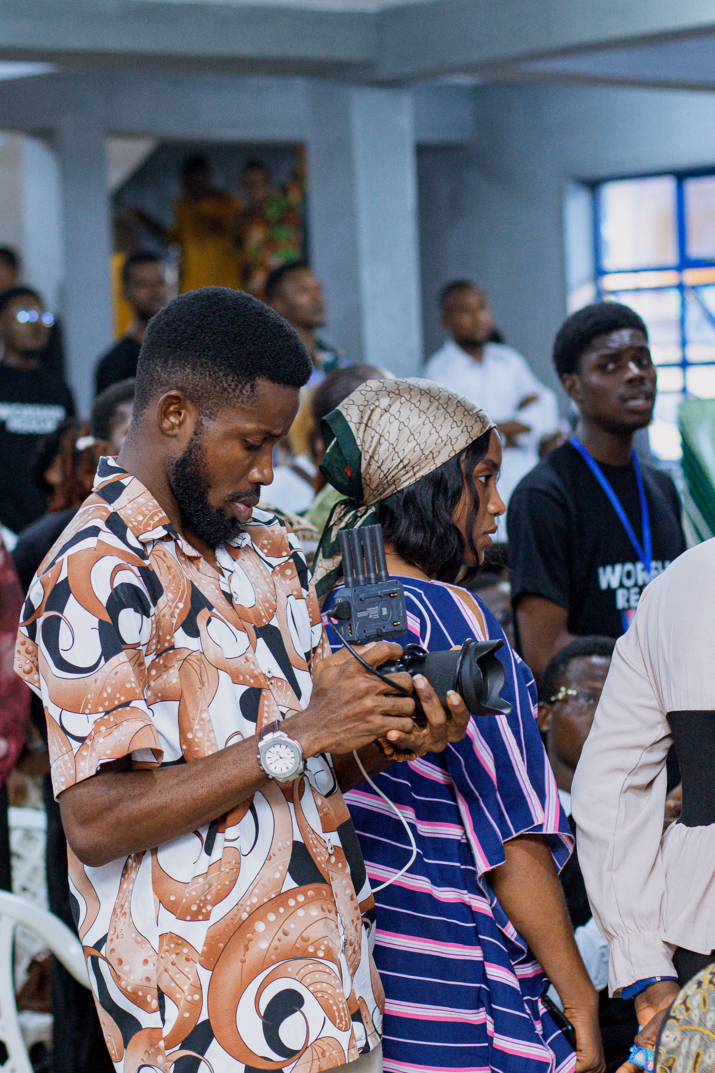 Camera operator in a patterned shirt filming from within a packed worship congregation