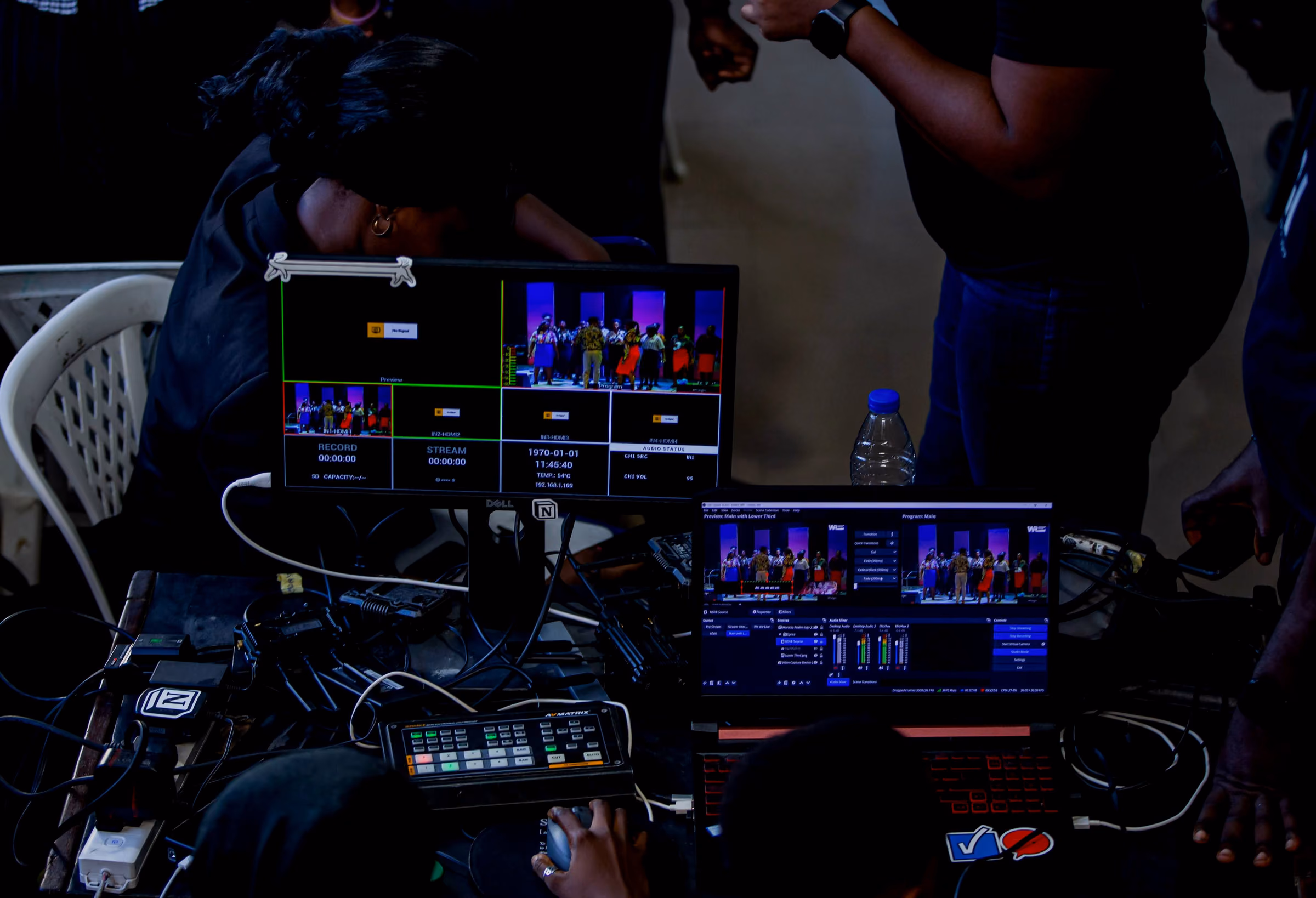 Two monitors showing switcher multiview and stage feeds above a tangle of control cables, in the dark